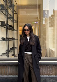 Long manteau noir par-dessus une chemise noire à boutons et un haut blanc, associé à un pantalon vert olive. Lunettes de soleil et étagères de chaussures noires en arrière-plan.