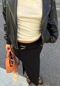 Zwarte rok met een zijsplit, aansluitend wit topje en zwart leren jacket. Houdt een oranje geweven handtas vast met een pluche charm. Zwarte sandalen.