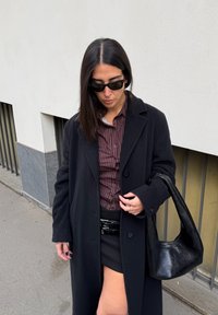 Czarny wełniany płaszcz, brązowa pasiasta koszula, czarna mini spódnica i czarna skórzana torba. Długie ciemne włosy, okulary przeciwsłoneczne w czarnych oprawkach. Betonowe tło.