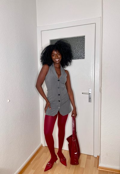 Chaleco gris sin mangas con detalles de botones, combinado con pantis rojos opacos y zapatos rojos de punta. Sosteniendo un bolso rojo a juego.