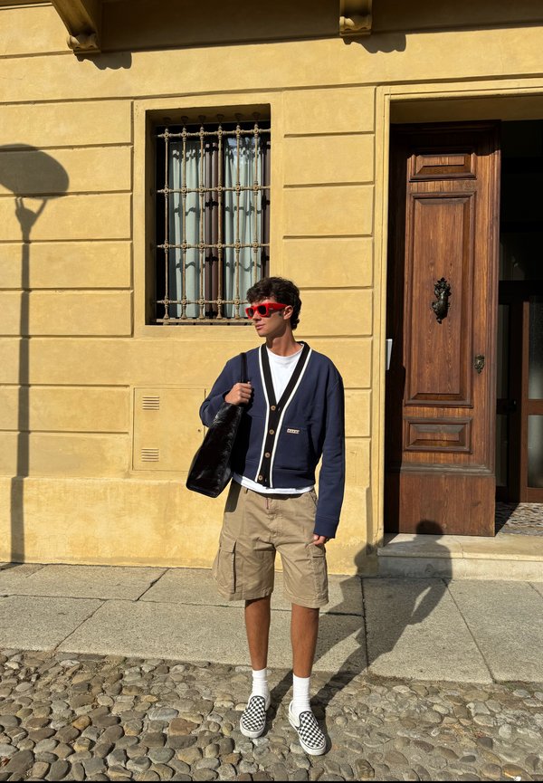 Námořnický kardigan s bílými akcenty a knoflíky, bílé tričko, béžové cargo šortky, černá taška přes rameno, kostkované slip-on boty, červené sluneční brýle.