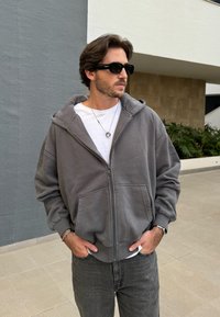 Hoodie cinza com fecho, feito de um tecido suave, apresentando um grande capuz e bolsos frontais. Usado por cima de uma camisa branca e jeans escuros. Óculos de sol pretos.