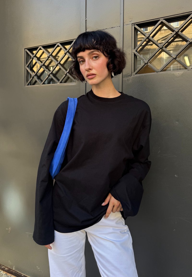 Jeune femme aux cheveux courts et foncés portant un haut à manches longues noir, un pantalon blanc et un sac à épaule bleu, debout devant une porte métallique grise.
