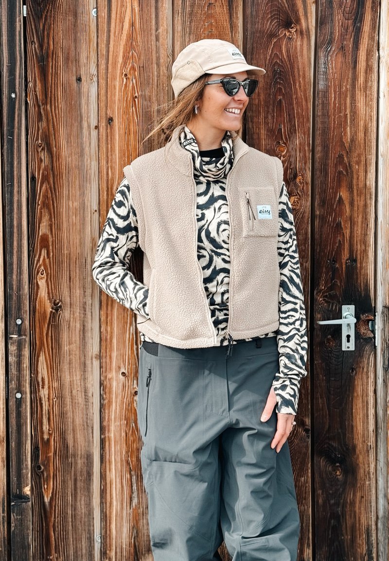 Woman wearing a beige cap, sunglasses, a patterned roll-neck jumper, a beige fleece waistcoat, and grey trousers standing against a wooden door, smiling.