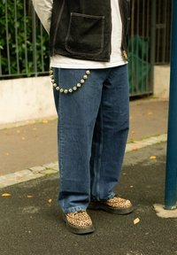 Denim jeans met een losse pasvorm, gecombineerd met schoenen met luipaardprint. Een zwarte vest met een voorzak en een decoratieve kralenketting op de tailleband.