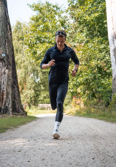 Hombre corriendo por un camino de grava, vistiendo una camiseta de manga larga negra y leggings negros. Zapatillas blancas y cinta para el sudor visibles. Árboles al fondo.