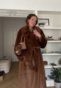 Femme portant un long manteau en fourrure marron, tenant un petit sac à main marron avec un porte-clés en forme d'ours en peluche, debout dans un salon moderne.