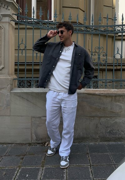 Hombre vistiendo una camisa gris de botones, camiseta blanca y pantalones de lino blancos. Zapatillas gris y blancas, de pie junto a una puerta decorativa.