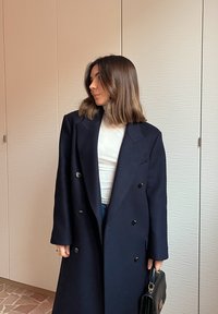 Femme aux cheveux bruns mi-longs portant un long manteau noir sur un haut blanc, tenant un sac à main noir, debout à l'intérieur.