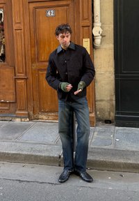 Άνδρας που φοράει μαύρο μπουφάν, πράσινα γάντια χωρίς δάχτυλα, γυαλιά ηλίου στο κεφάλι και τζιν, ρυθμίζει τα γάντια του ενώ στέκεται σε πεζοδρόμιο πόλης κοντά στην πόρτα αριθμός 42.