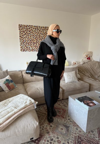 Mujer con vestido negro, pañuelo beige en la cabeza, suéter gris atado alrededor de los hombros, botas negras, sosteniendo un bolso negro en una sala de estar beige.