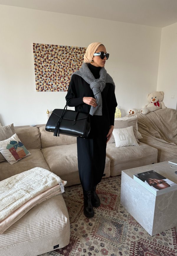 Femme en robe noire, foulard beige sur la tête, pull gris noué autour des épaules, bottes noires, tenant un sac à main noir dans un salon beige.