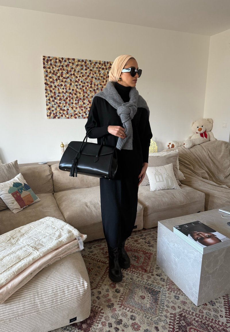 Femme en robe noire, foulard beige sur la tête, pull gris noué autour des épaules, bottes noires, tenant un sac à main noir dans un salon beige.