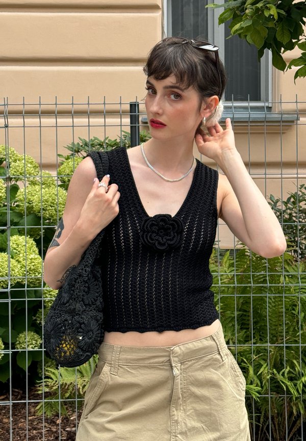 Débardeur en crochet noir avec un col en V et un détail floral, associé à un short beige et un sac en crochet assorti. Feuillage vert en arrière-plan.