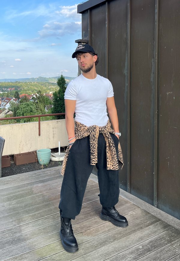 Ung mand iført hvid t-shirt, sort kasket, sorte vide bukser, sorte støvler og en leopardprintjakke bundet om taljen står på en træterrasse.