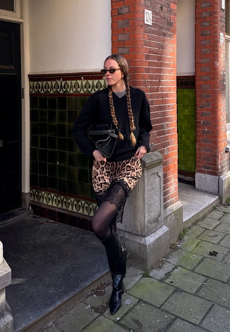 Femme aux cheveux tressés portant des lunettes de soleil, un pull noir, une jupe imprimée léopard, des collants noirs et des bottes, appuyée contre un coin de bâtiment en briques carrelées.