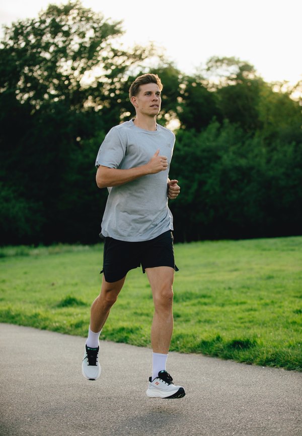 Corredor masculino atlético con una camiseta gris claro y pantalones cortos negros, vistiendo zapatillas de correr blancas y negras con calcetines blancos, en un camino pavimentado.