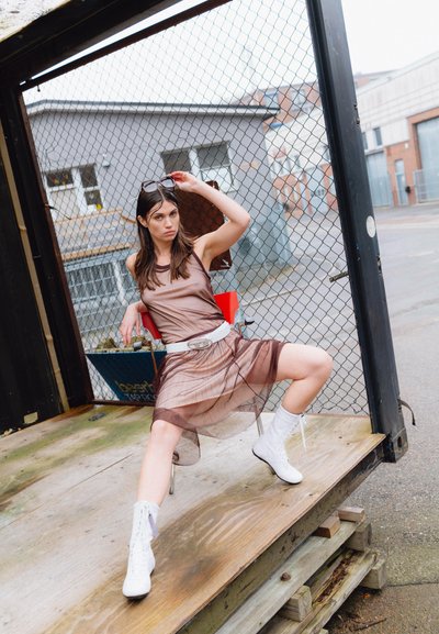 Vestido marrón transparente con un corsé ajustado y una falda fluida, combinado con zapatillas altas blancas. La modelo posa sobre una plataforma de madera con un fondo de valla.