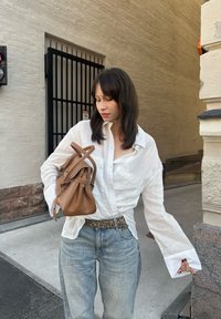 Witte lange mouwen shirt met knoopmanchetten, lichtblauwe spijkerbroek en een bruine leren tas. Geweven achtergrond met subtiele lijnen en een donkere poort.