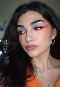 Kvinna med långt mörkt hår som bär färgglad ögonsmink med gröna, blå och röda nyanser, kombinerat med guldörhängen och ett guldhalsband.