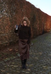 Femme portant des lunettes, un cardigan noir et une jupe marron, tenant une tasse de café en marchant sur un chemin pavé à côté d'une haute haie d'automne.