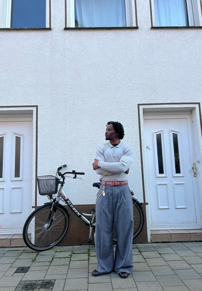 Una persona está de pie junto a una bicicleta en un pavimento adoquinado, vistiendo un suéter gris de canalé y pantalones holgados a rayas con un cinturón estampado.