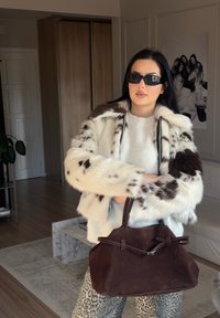 Giacca in pelliccia sintetica bianca e marrone con colletto alto, abbinata a un maglione leggero. Tiene una borsa in suede marrone con un dettaglio a nodo.