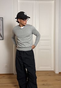 Moški, ki nosi črno baseball kapo, očala, sivi pulover in črne cargo hlače, stoji v notranjosti z rokami v žepih, obrnjeno levo.