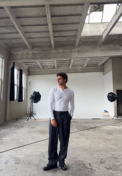 Modelo con una camisa henley blanca de manga larga y pantalones negros, de pie en un estudio industrial espacioso con grandes ventanas y luces softbox.