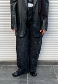 Czarne szerokie spodnie z denimu ozdobione okrągłymi srebrnymi przelotkami. Noszone z czarną skórzaną marynarką i błyszczącymi czarnymi butami.