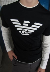 Čierne tričko s krátkym rukávom a bielym logom, na ktorom je štýlizovaný orol a text "GA", nosené cez biele tričko s dlhým rukávom.