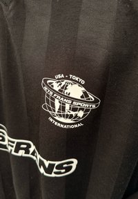 Czarny sportowy shirt w paski z białym logo globu. Tekst zawiera "USA - TOKYO" oraz "JETS FRANS SPORTS INTERNATIONAL." Miękka faktura.