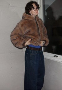 Persona con gafas, vistiendo una chaqueta marrón esponjosa y jeans azul oscuro, ajustando el dobladillo de la chaqueta cerca de una ventana con una pared asegurada con cinta adhesiva detrás.