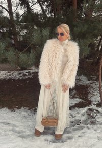 Chaqueta de piel sintética color crema, vestido blanco con encaje en capas, bolso marrón claro y botas con plataforma marrón; ubicados en un área exterior nevada con pinos.