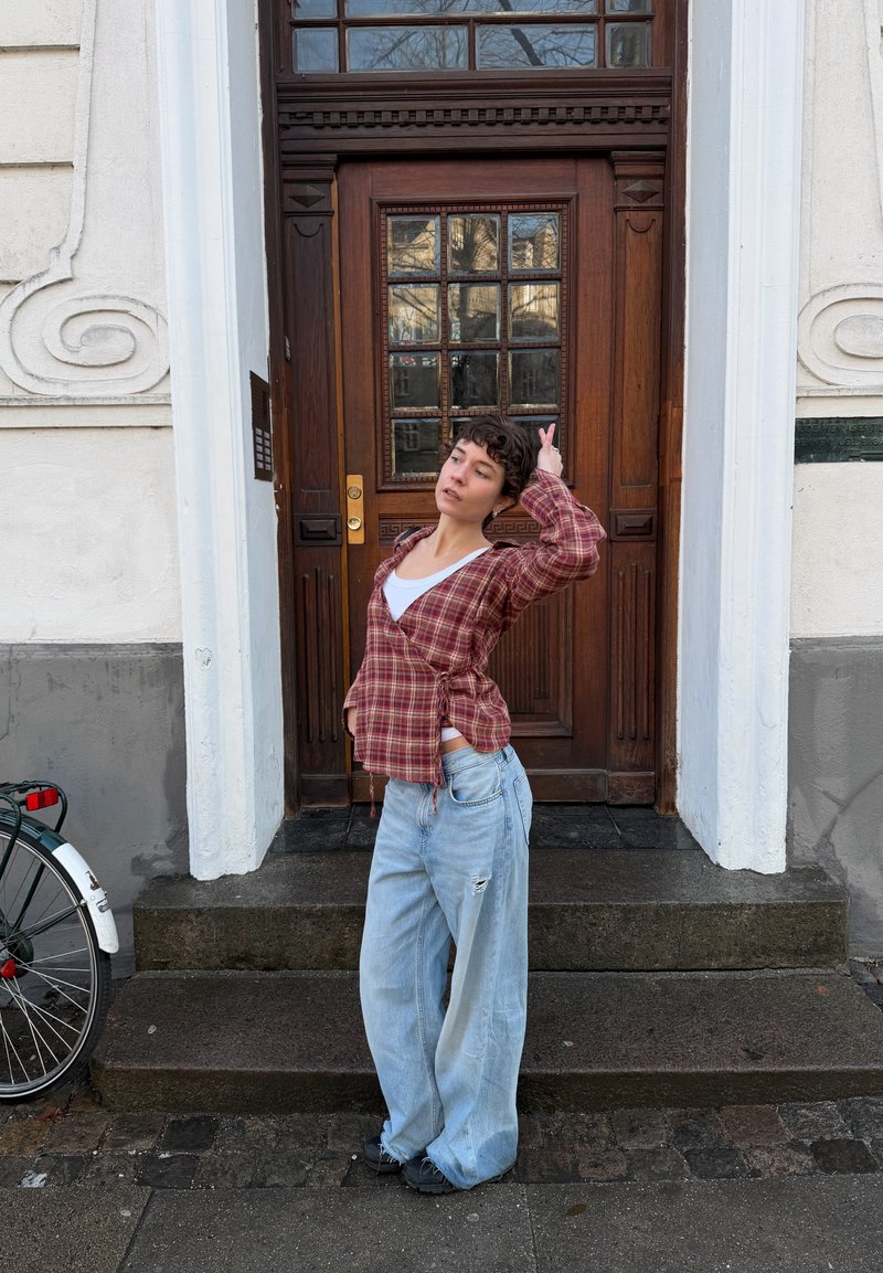 Jeune personne portant une chemise à carreaux et un jean ample, posant devant une porte en bois avec des vitres, à côté d'une roue de bicyclette.