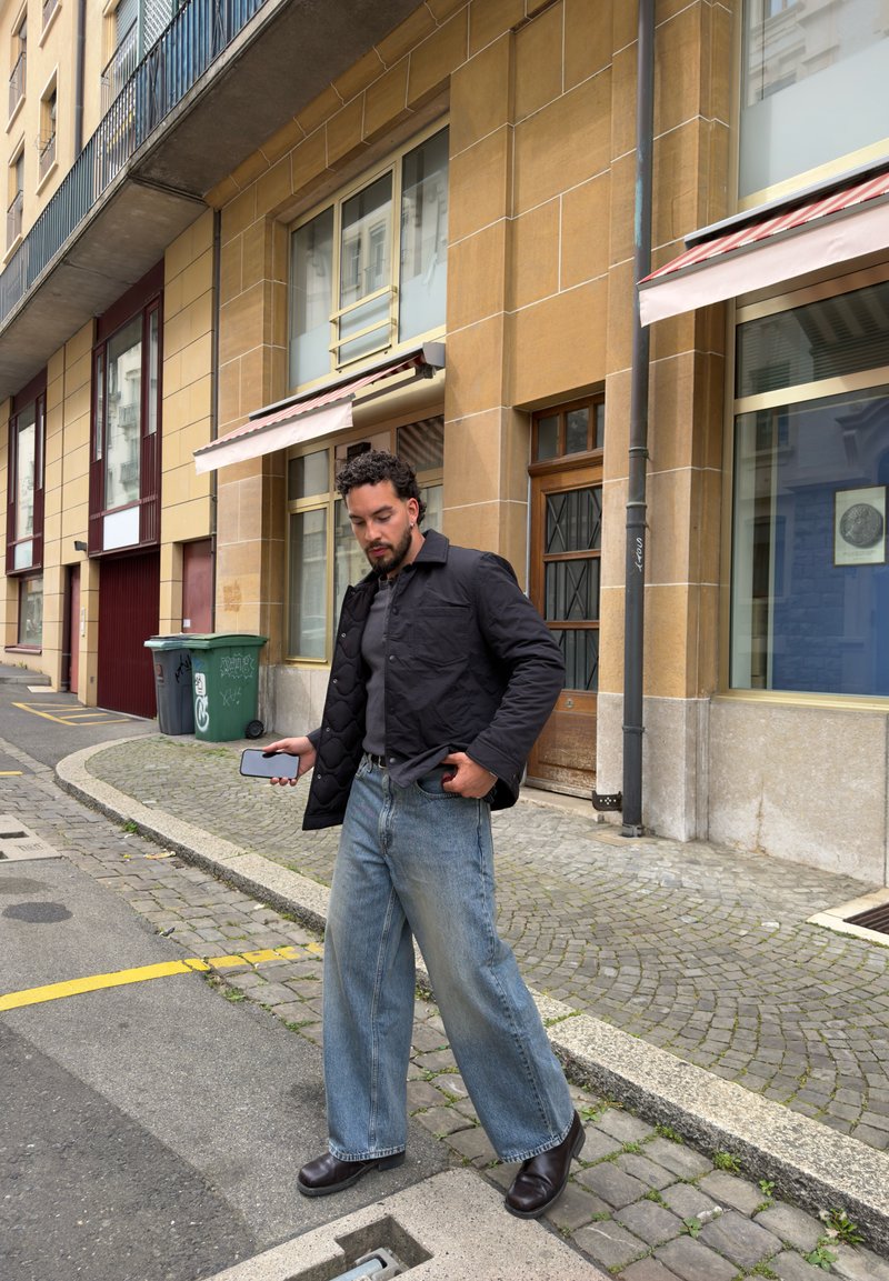 Homme en veste noire et jeans amples tenant un smartphone, marchant dans une rue pavée à côté de bâtiments avec de grandes fenêtres et des poubelles vertes.