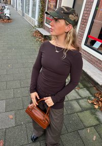 Femme portant une casquette camouflage, un haut à manches longues foncé et un pantalon ample, tenant un sac à main marron sur un trottoir en ville, avec des vélos et des feuilles tombées à proximité.