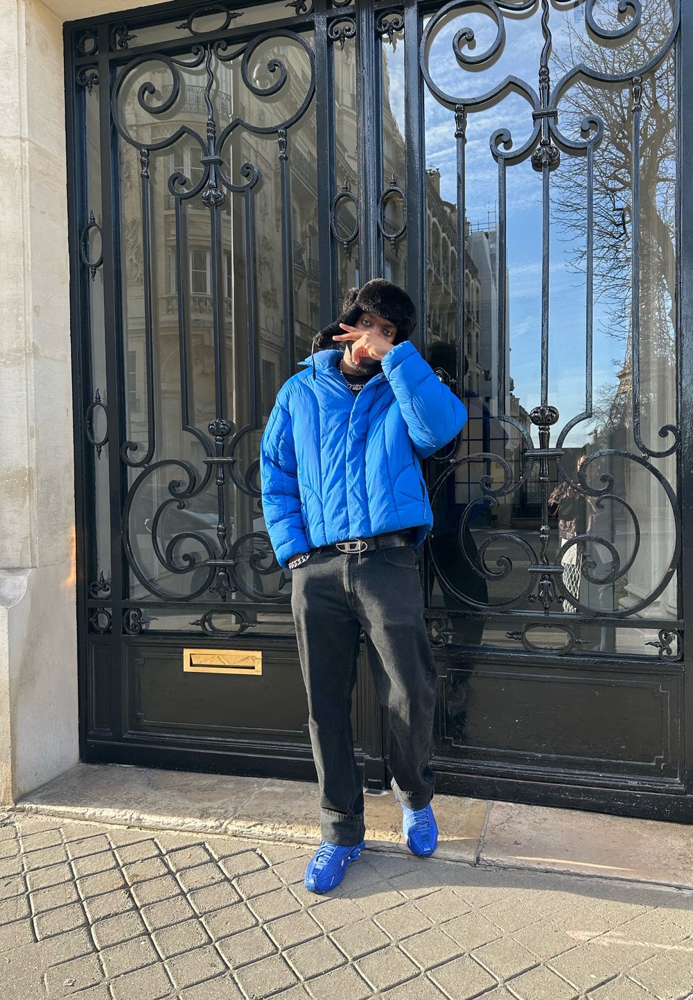 Blå polstret jakke, sorte bukser, blå sneakers. Står ved en dekorativ sort metalport. Blød tekstur og markant farvekontrast.