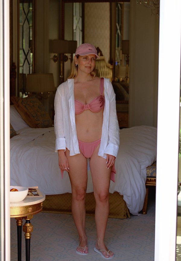 Vrouw staand binnenshuis, gekleed in een roze bikini, wit open shirt, roze pet en slippers, met op de achtergrond een bed en sierlijk meubilair.