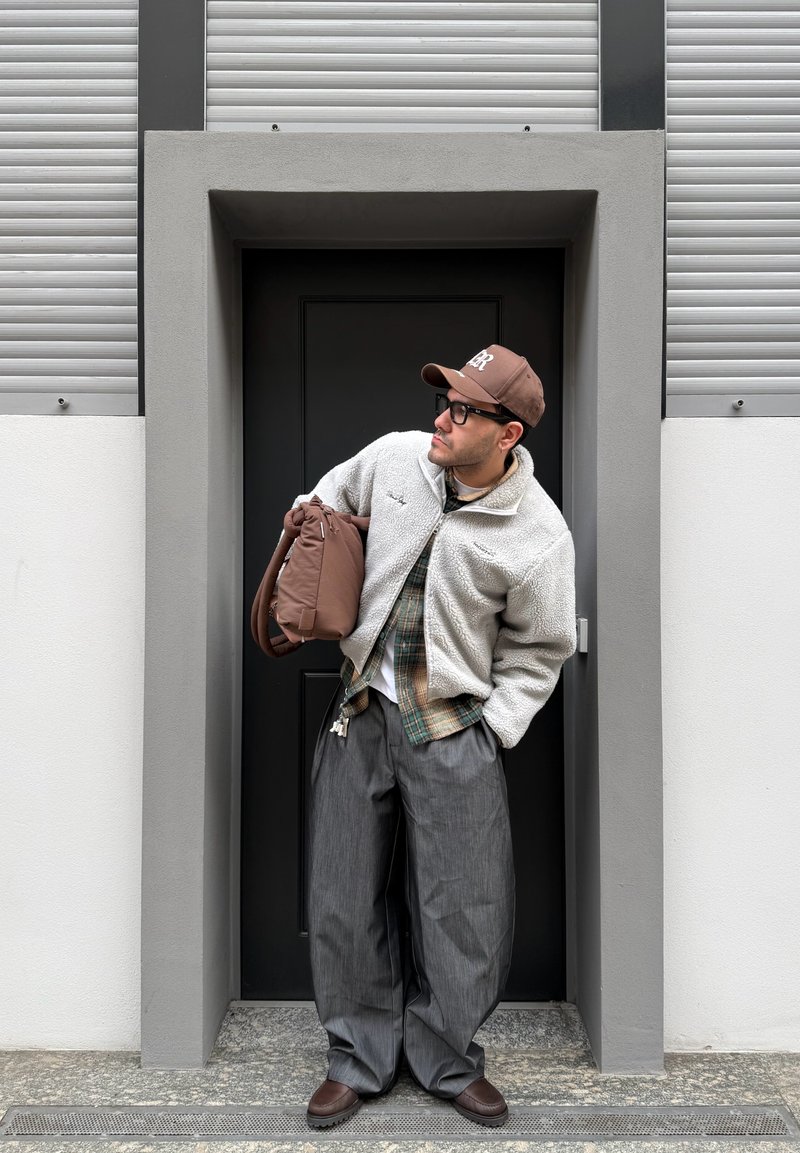 Homme portant des lunettes, casquette marron, veste polaire blanche, chemise à carreaux, pantalons larges gris, tenant un sac marron, debout dans une porte noire en retrait.