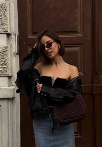Veste en cuir noire portée sur un haut noir sans bretelles, jupe en jean bleue et un sac à main tressé rouge foncé. De petites lunettes de soleil rondes complètent la tenue.