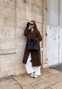 Femme portant un manteau marron à carreaux, un pantalon blanc large, des gants noirs et des lunettes de soleil, tenant un sac à main noir, debout contre un mur en pierre en plein air.