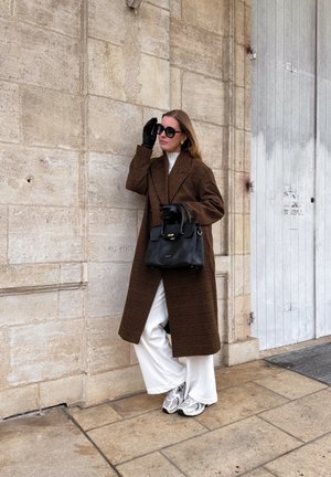 Mujer con abrigo de cuadros marrones, pantalones anchos blancos, guantes negros y gafas de sol, sosteniendo un bolso negro, de pie contra una pared de piedra al aire libre.