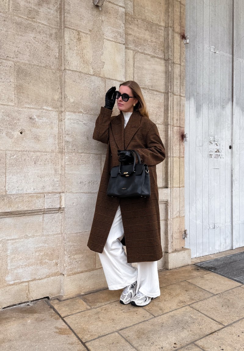 Femme portant un manteau marron à carreaux, un pantalon blanc large, des gants noirs et des lunettes de soleil, tenant un sac à main noir, debout contre un mur en pierre en plein air.
