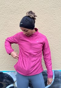 Rózsaszín könnyű dzseki rugalmas derék- és mandzsettaszegéllyel, cipzáras záródással és Nike logóval. Szürke leggings fölött viselve, texturált bézs fal előtt.