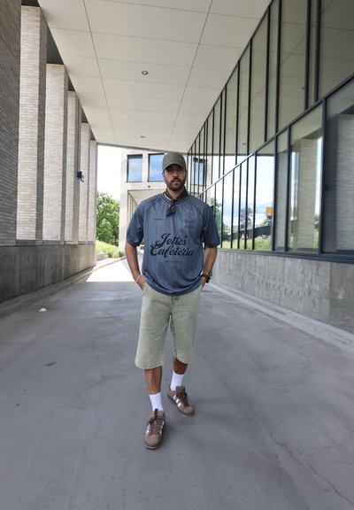 Jersey atlético gris con acentos y letras oscuros, pantalones cortos verde claro, zapatillas marrones y una gorra verde. De pie en un corredor con grandes ventanales.