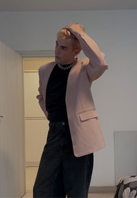Lyserød blazer med hakkede revers, båret over en sort skjorte. Sorte bukser med høj talje, glat tekstur og minimal synlig hardware.