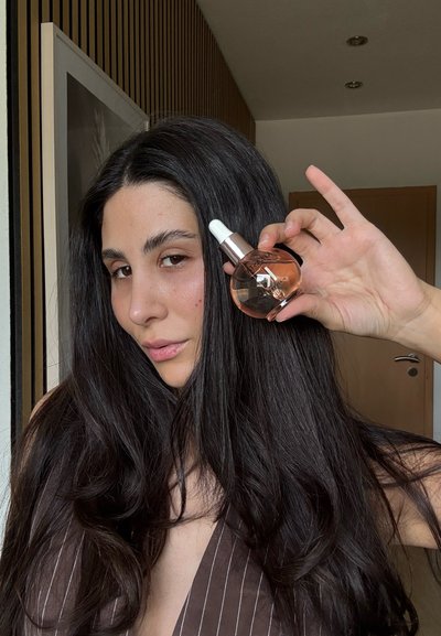 Botella transparente con un líquido de color rosa-naranja, cuentagotas blanco y tapa metálica. Pelo largo y oscuro, usando una blusa de rayas marrón, con el hombro visible.