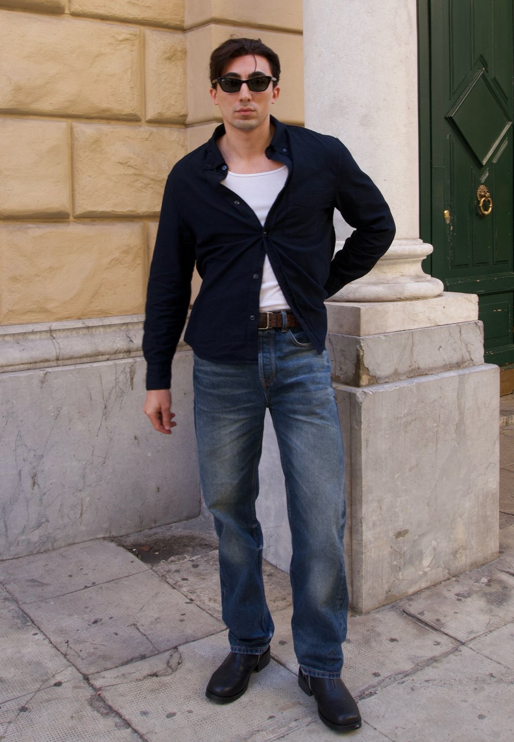 Schwarzes Hemd mit Knöpfen, weißes Tanktop, verwaschene blaue Jeans, schwarze Lederschuhe und Sonnenbrille, posiert neben einer Steinsäule und einer grünen Tür.