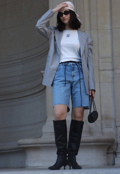 Blazer gris, camiseta blanca con logo, pantalones cortos de mezclilla azul, botas negras de tacón alto hasta la rodilla, sosteniendo un pequeño bolso negro, sobre un fondo neutro.